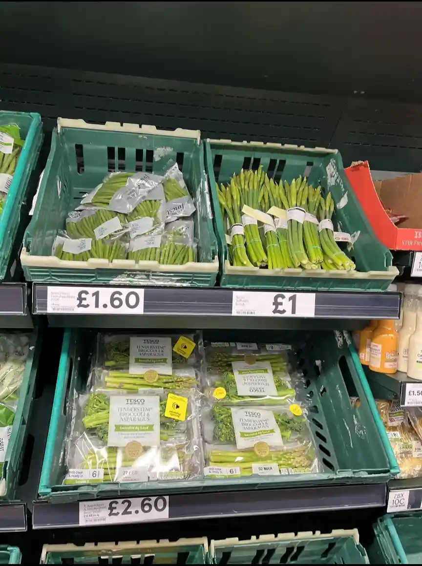A supermarket produce aisle shows green crates of vegetables with price tags: £1.60 for extra fine beans, £1 for daffodil…