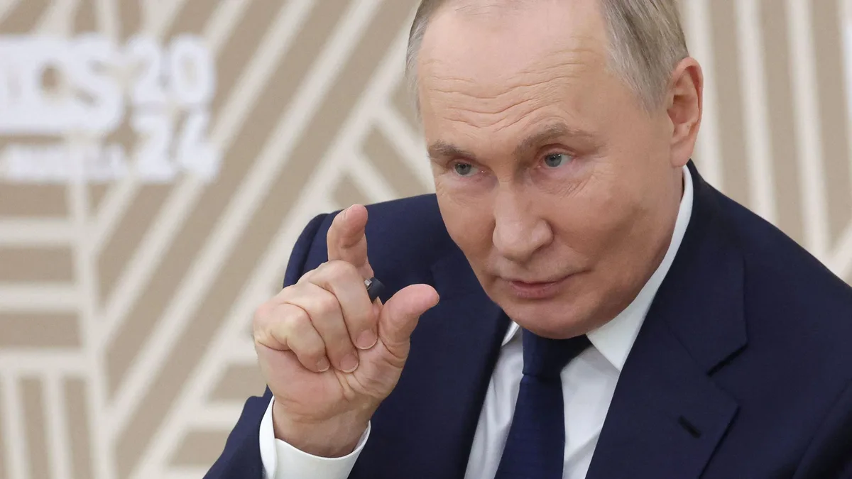 A close-up photo of Vladimir Putin in a dark suit and tie, pointing his index finger forward with a serious expression. The…