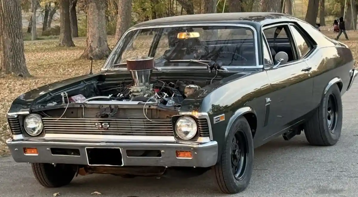 A black classic Chevrolet SS muscle car with its hood open, revealing a heavily modified engine with a large chrome air…