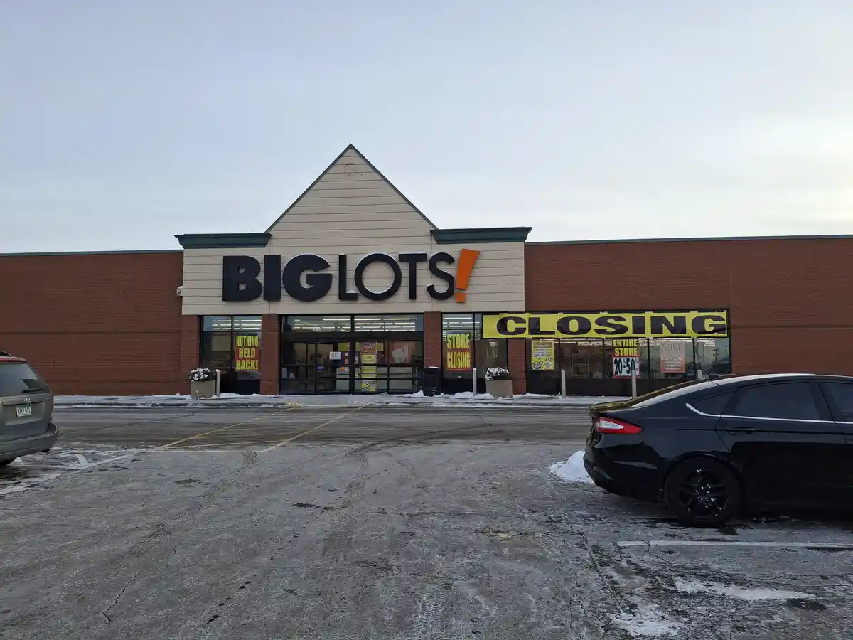 An exterior photo of a Big Lots retail store with a large yellow banner across the front windows reading 'CLOSING' and…