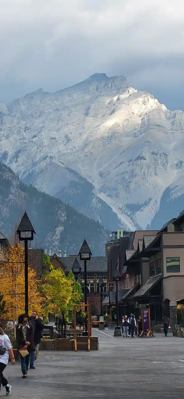 A scenic photo of a pedestrian street in a mountain town with large, snow-dusted peaks in the background and autumn-colored…