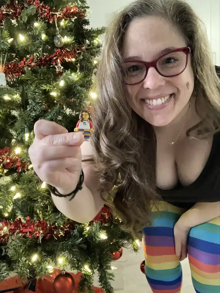 A smiling woman with curly hair and red glasses holds up a small Lego minifigure in front of a decorated Christmas tree with…