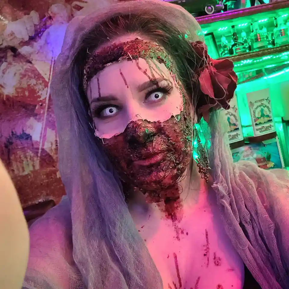 A person in a wedding veil and dress is shown with gruesome zombie makeup, including fake blood, torn flesh, and glittery…