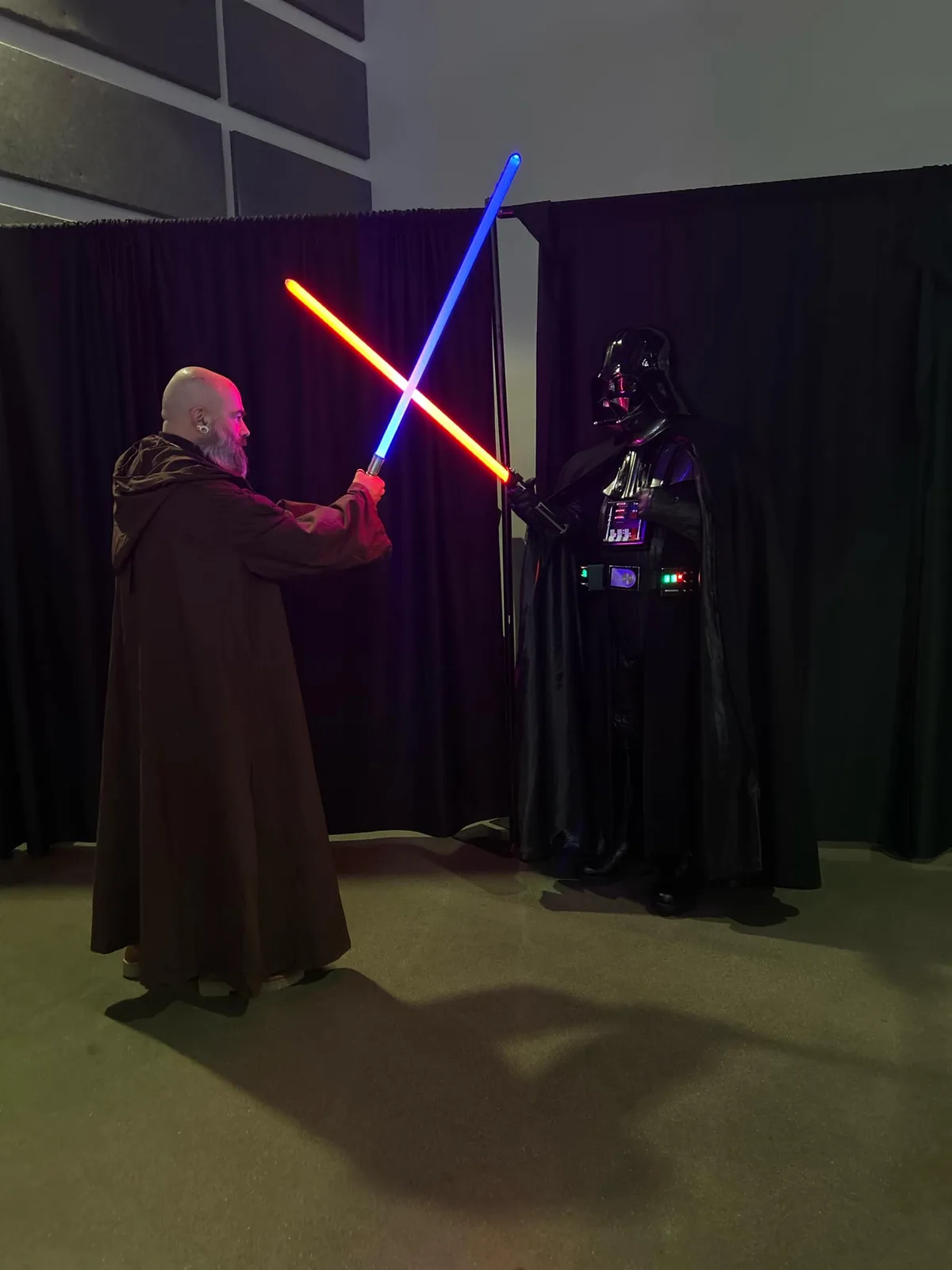 Two people in Star Wars costumes stand facing each other in a dark room with black curtains, holding crossed lightsabers—one…
