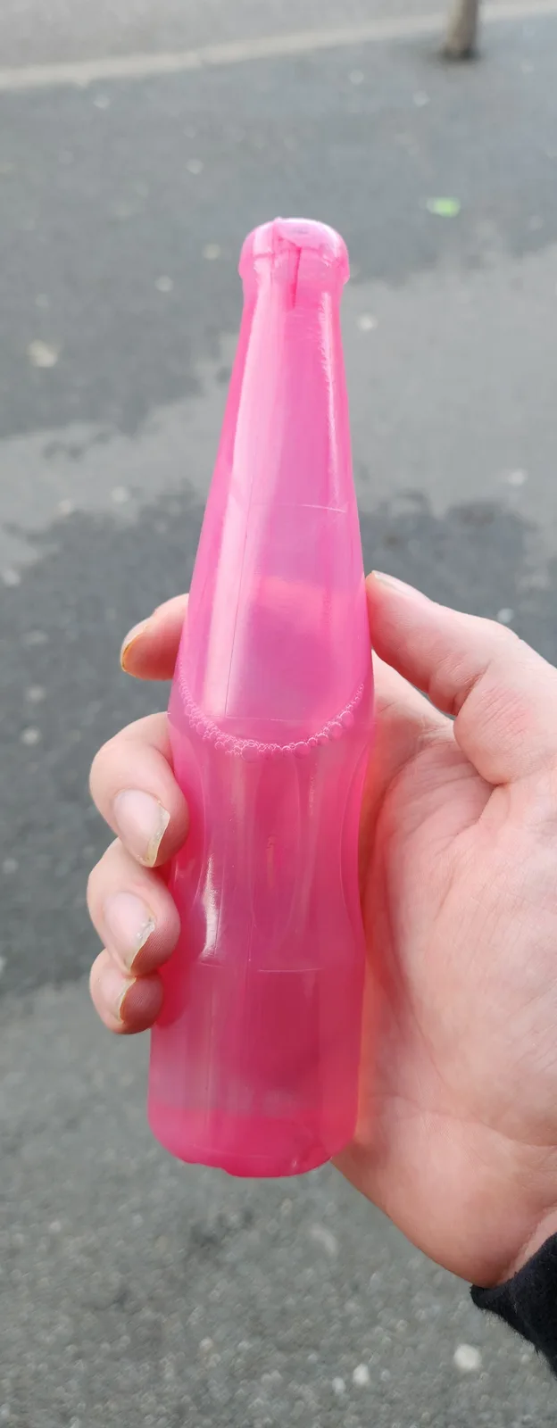 A hand holds a bright pink plastic bottle filled with pink liquid, with a bubble ring visible near the top. The background…