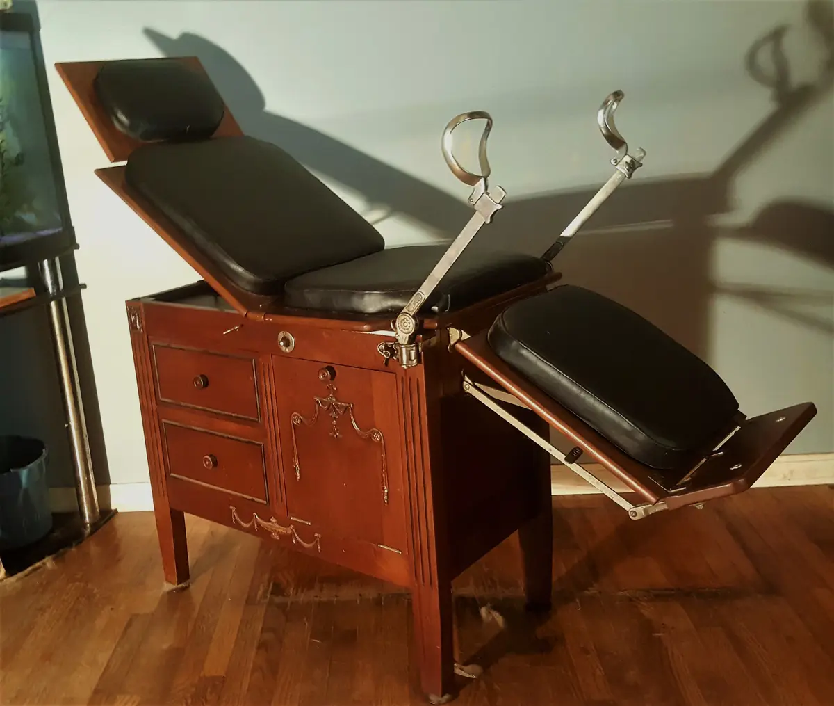 An antique wooden gynecological examination table with black leather cushions and metal stirrups is positioned in a room…