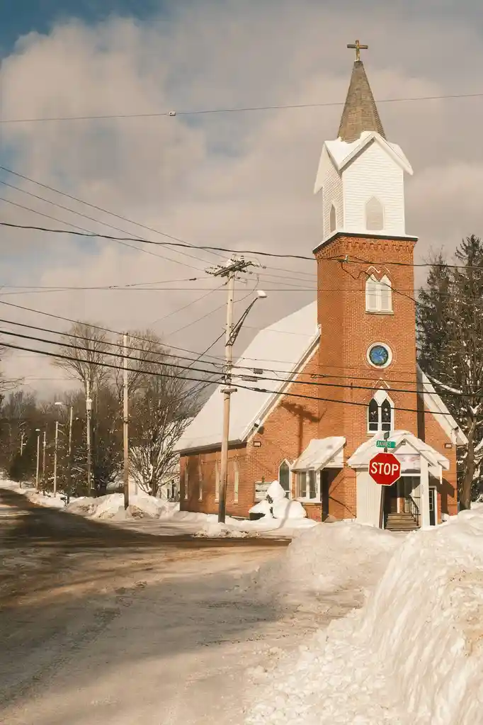 A snowy street scene features a red brick church with a white steeple topped by a cross, surrounded by snowbanks and utility…