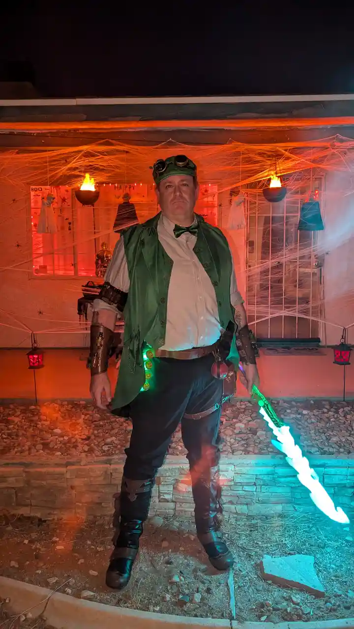 A man in a steampunk-style Halloween costume stands outside a house decorated with orange lights, spiderwebs, and ghost…