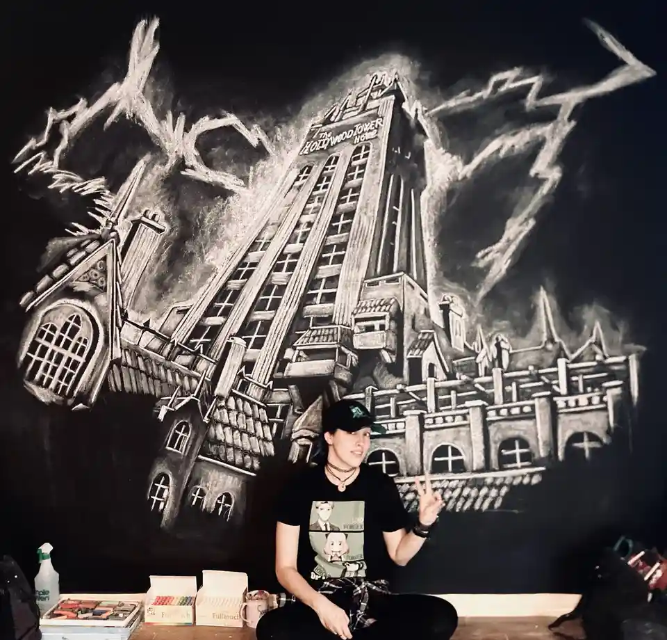 A person sits in front of a large chalk drawing of the Hollywood Tower Hotel, a famous haunted attraction from Disney's…