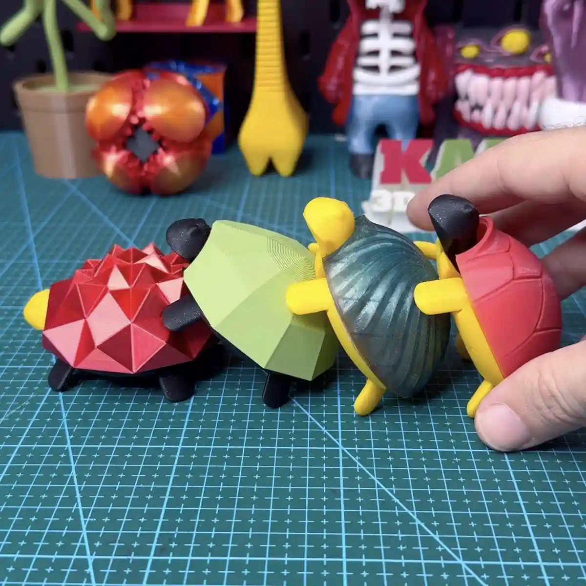 A hand holds a red 3d-printed turtle figurine with a smooth, rounded shell, next to three other 3d-printed turtle figurines…