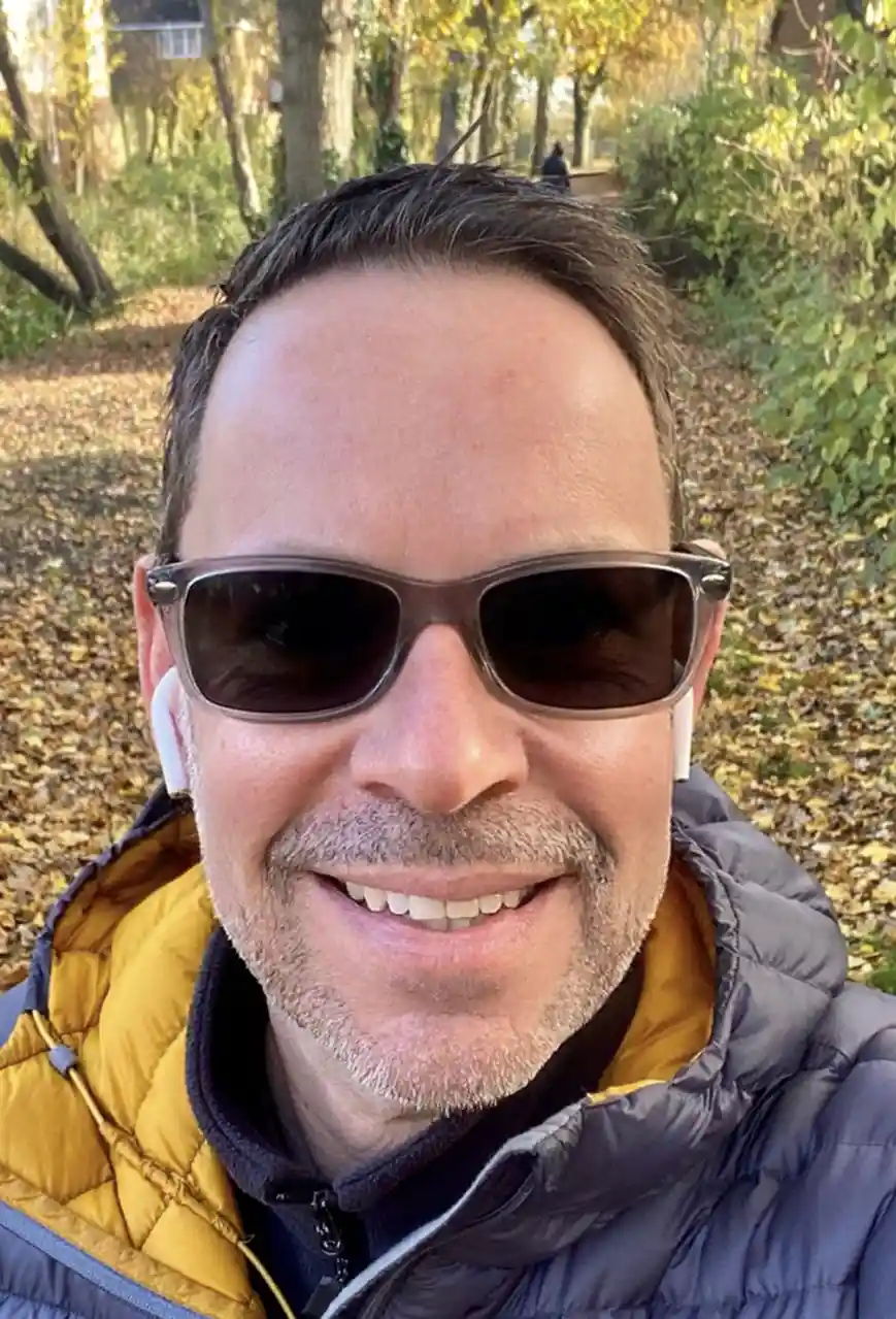A man with a gray beard and short brown hair smiles at the camera while wearing dark sunglasses and white earbuds. He is…