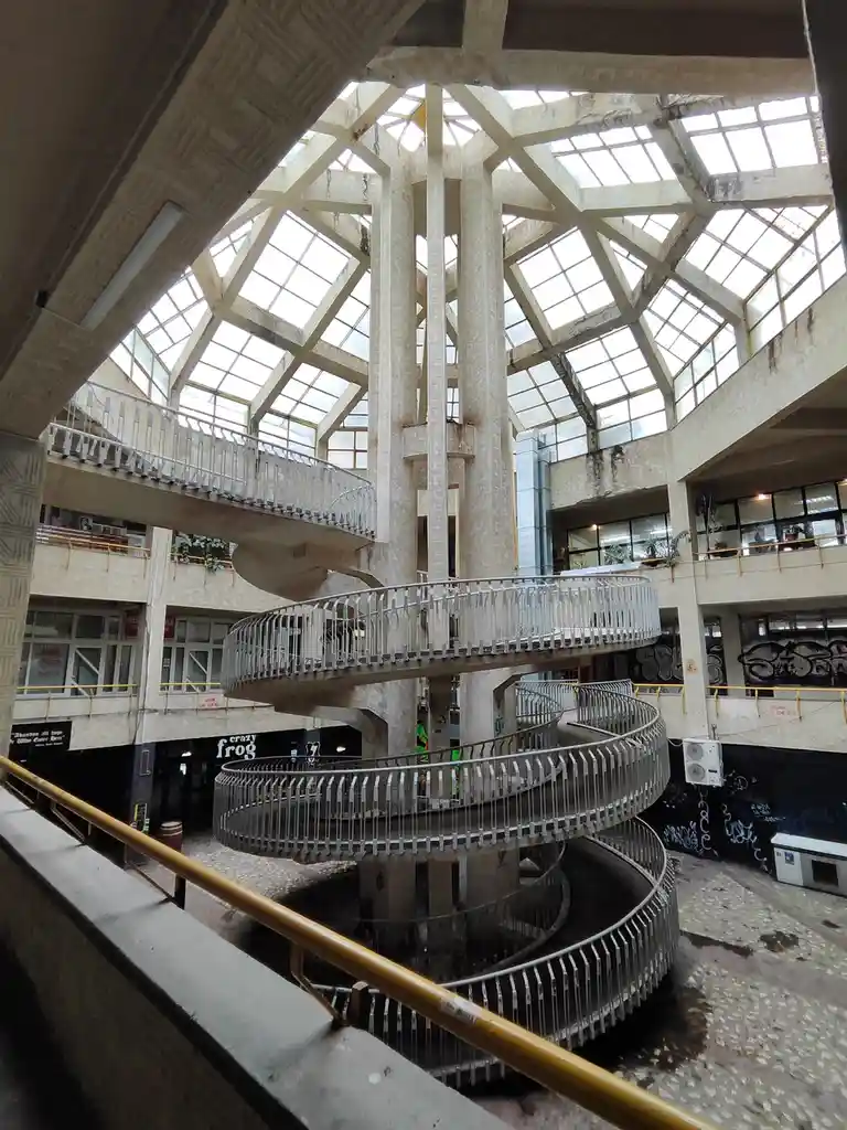 An upward-looking shot of a spiraling, multi-level concrete and metal walkway inside a decaying, abandoned building with a…