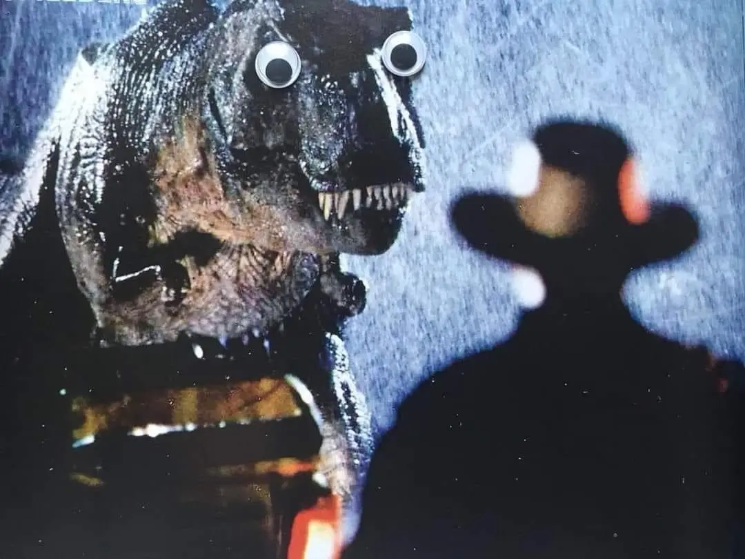 A close-up image shows a large, textured, dark-colored dinosaur puppet with googly eyes, facing a blurry silhouette of a…