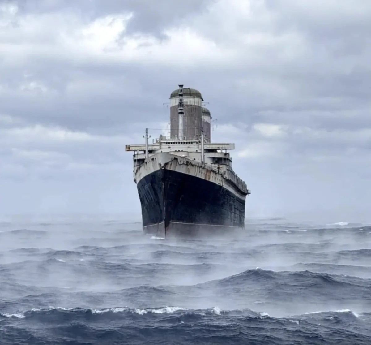 A large, weathered ship with a dark hull and white superstructure is shown stern-on, battling rough, choppy seas under a…