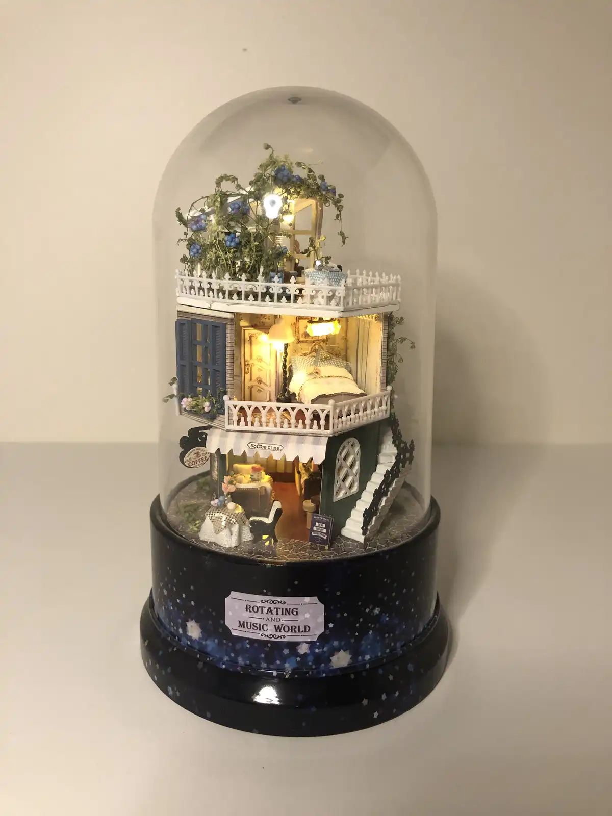A miniature, multi-level dollhouse under a glass dome, featuring a cozy bedroom upstairs and a coffee shop downstairs, with…