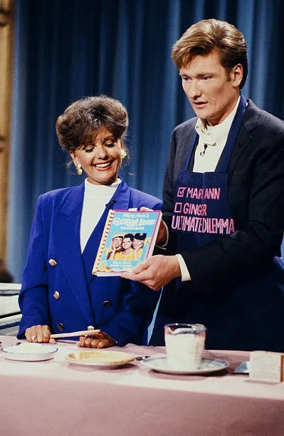 A man and woman stand at a cooking show set, the man wearing an apron with the text 'I'M MARIANN I'M GINGER ULTIMATEDILEMMA'…