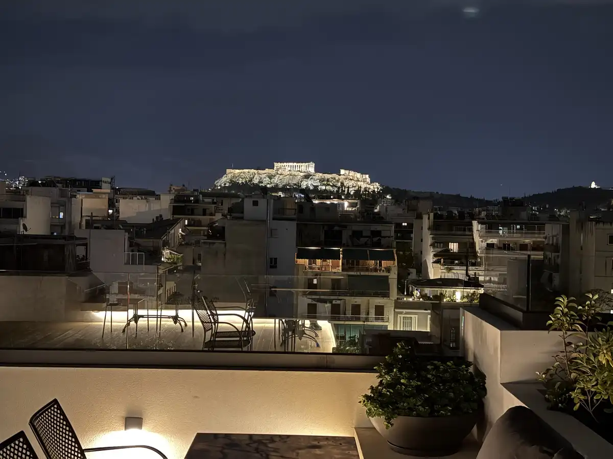 A nighttime photo from a rooftop terrace shows a view of the illuminated Acropolis of Athens in the distance, surrounded by…