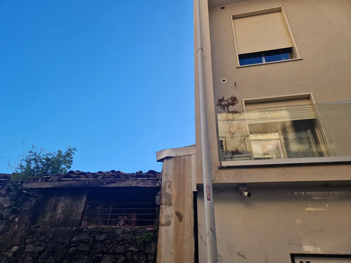 A modern beige building with glass balconies and security cameras stands beside a dilapidated stone structure with a rusted…