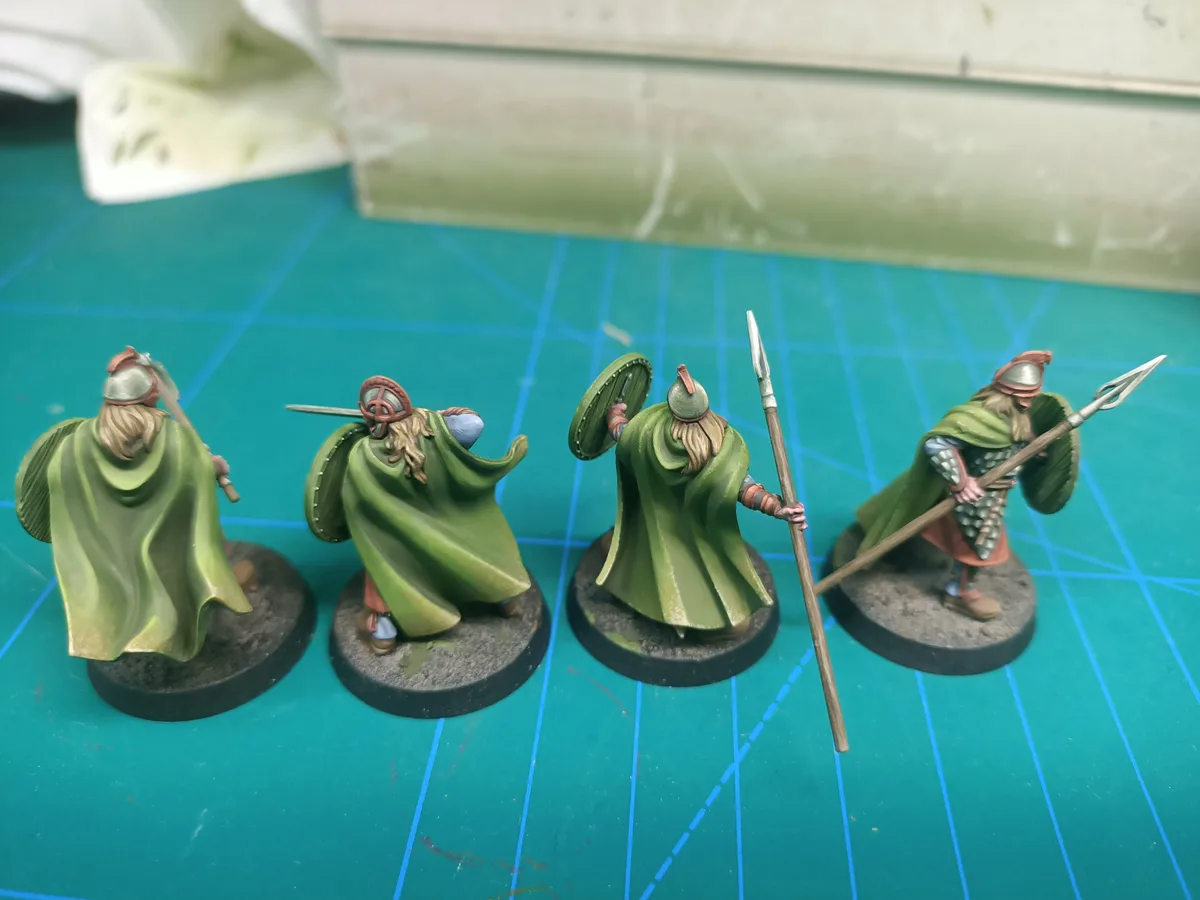 Four painted miniature fantasy warriors in green cloaks stand in a row on a green grid mat, each holding different weapons…