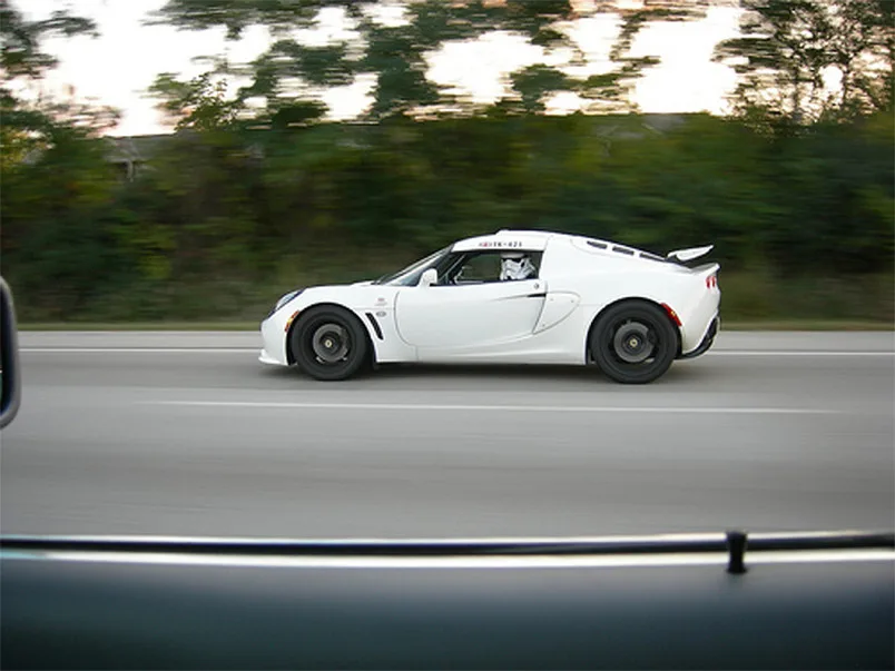 A white sports car, likely a Lotus Elise, is captured in motion on a road with blurred background trees, viewed from inside…