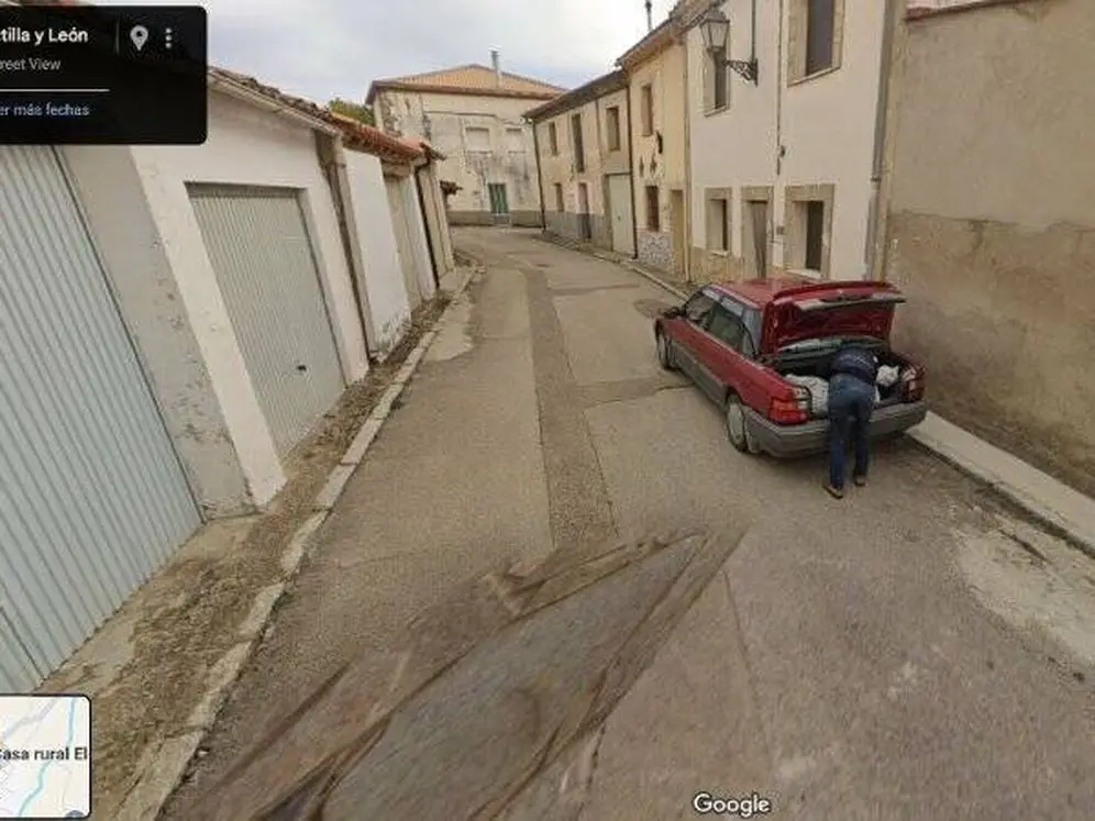A Google Street View image shows a person bent over the open trunk of a red car parked in a narrow, dead-end street. The car…