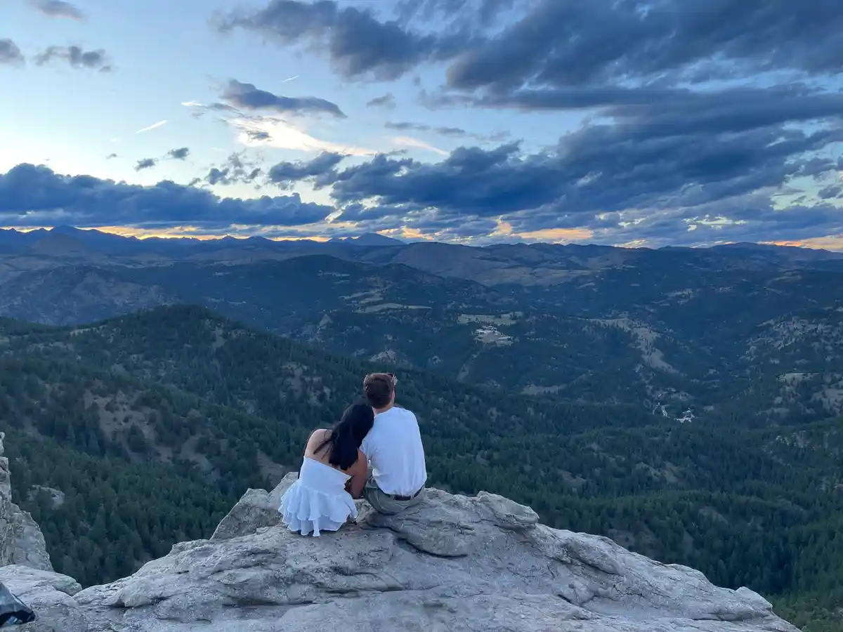 A couple sits side by side on a rocky mountain peak, backs to the camera, overlooking a vast valley with rolling hills and…