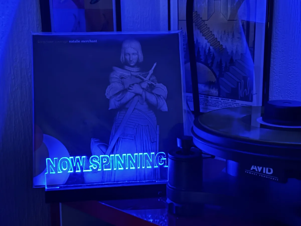 A vinyl record sleeve for Natalie Merchant's 'Keep Your Courage' is displayed next to a turntable under blue lighting, with…