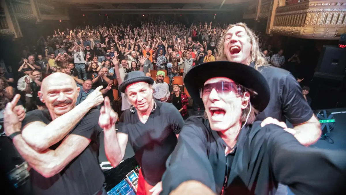 Three men, likely musicians, are on a stage in front of a large, cheering crowd. The man in the foreground wears a black hat…