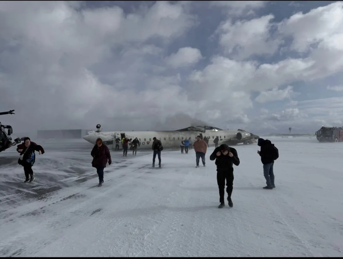 A snowy airport scene shows passengers evacuating a damaged airplane with smoke rising from its fuselage. Several people are…