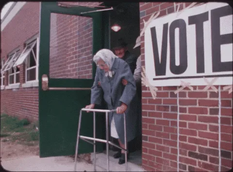 A sequence of frames shows an elderly woman in a blue coat and white headscarf using a metal walker to enter a brick…