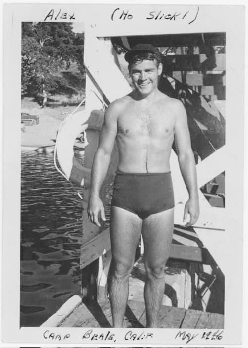 A vintage black-and-white photo shows a smiling young man in swim trunks and a cap standing on a wooden dock by water…