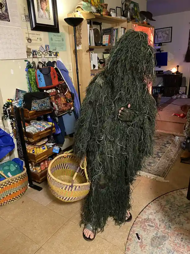 A person wearing a full-body green ghillie suit stands indoors in a cluttered room, holding a woven basket. The suit is made…