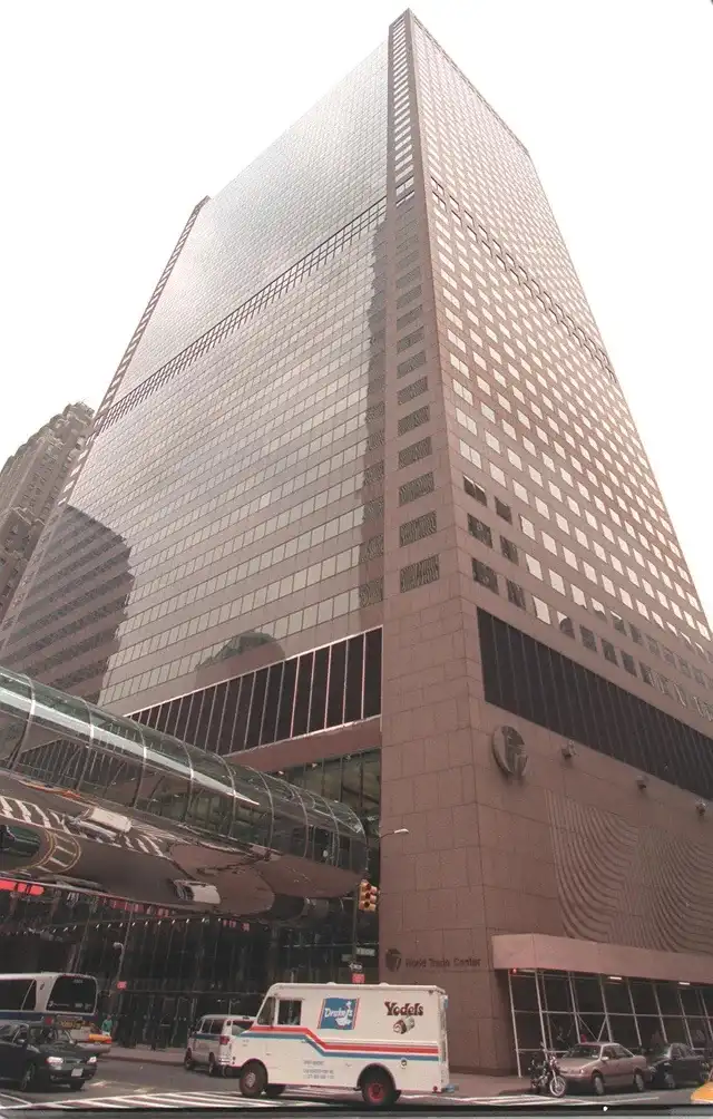 A low-angle photo of the World Trade Center in New York City, showing its glass and stone facade with a skywalk and…
