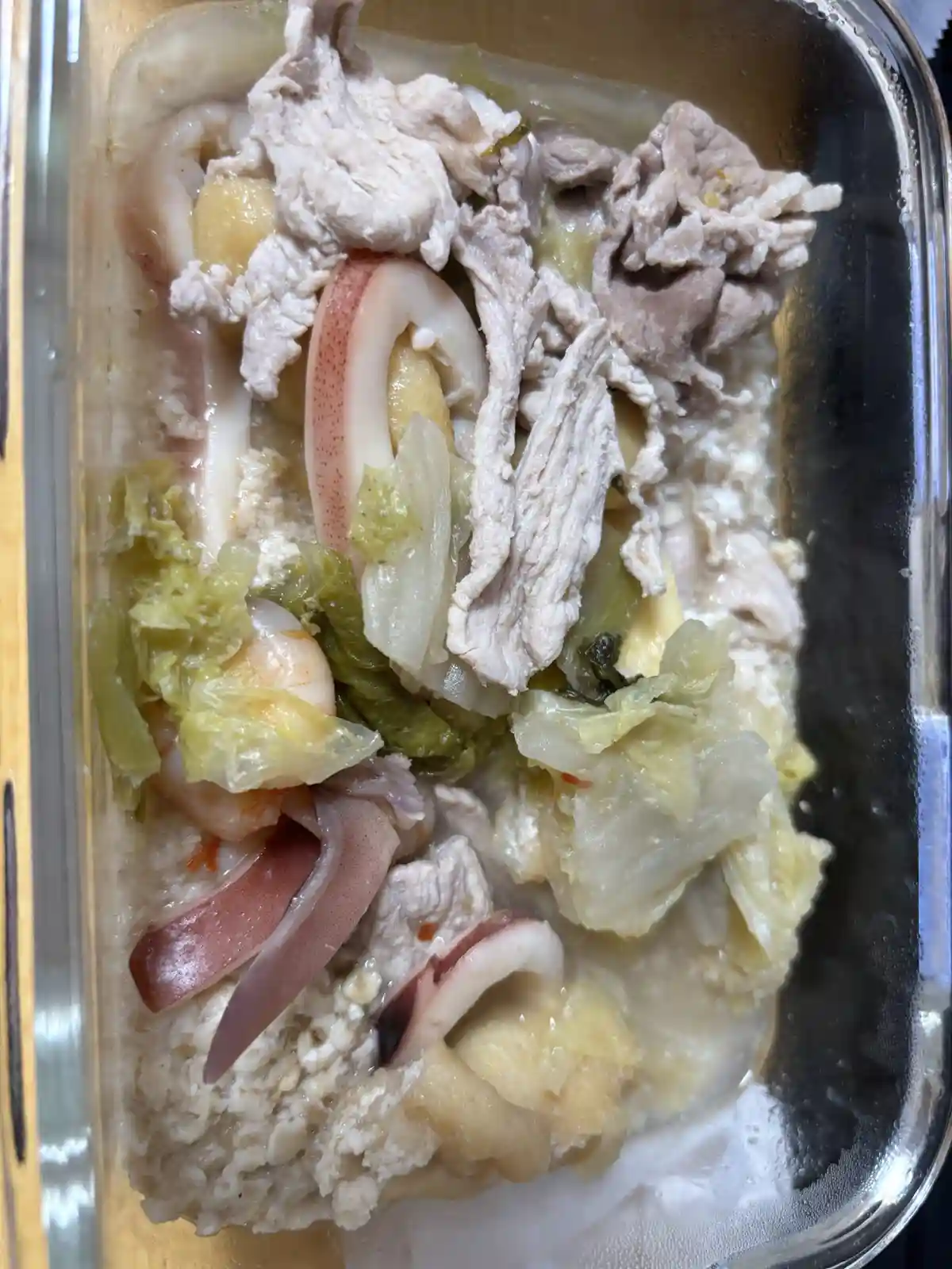 A top-down photo of a glass container filled with a mixed meal including shredded meat, sliced squid, shrimp, pickled…