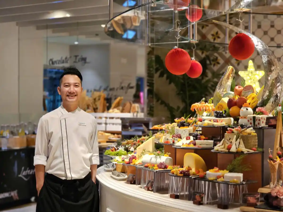 A smiling man in a white chef's coat stands in front of an elaborate, multi-tiered buffet display filled with various…