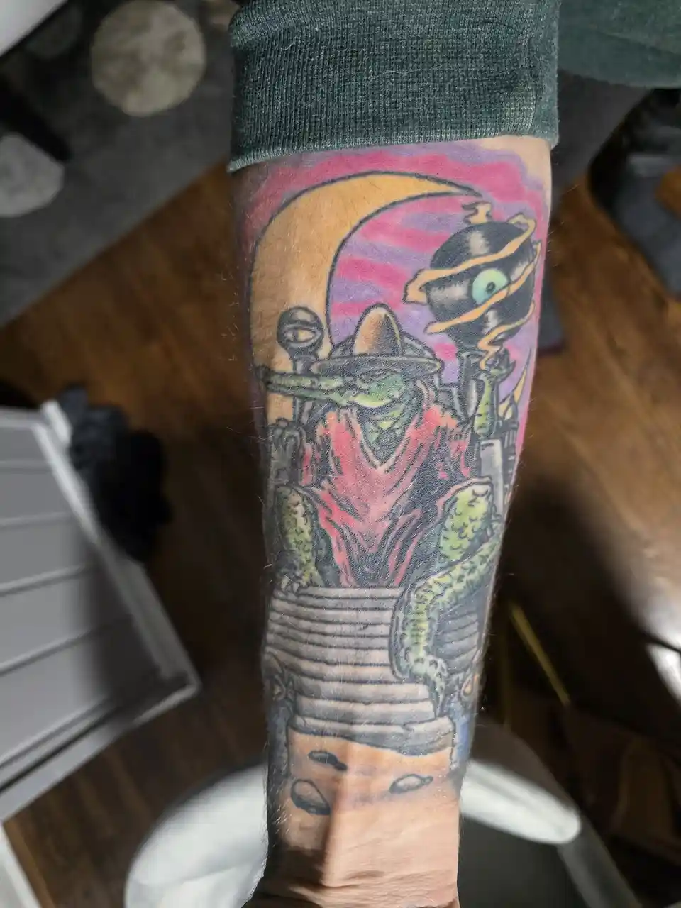 A person's forearm displays a colorful tattoo featuring characters from the movie 'The Thing,' including a green, reptilian…