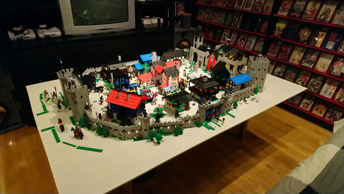 A large, intricate Lego castle and town model sits on a white table, surrounded by green Lego grass and trees, with tiny…