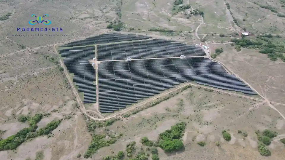 An aerial drone photo shows a large, organized field of solar panels arranged in neat rows across a dry, hilly, grassy…