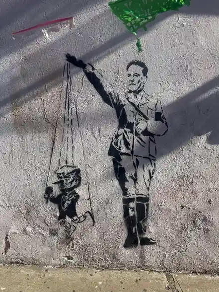 A black stencil graffiti on a textured white wall depicts a man in a military-style uniform holding strings that control a…