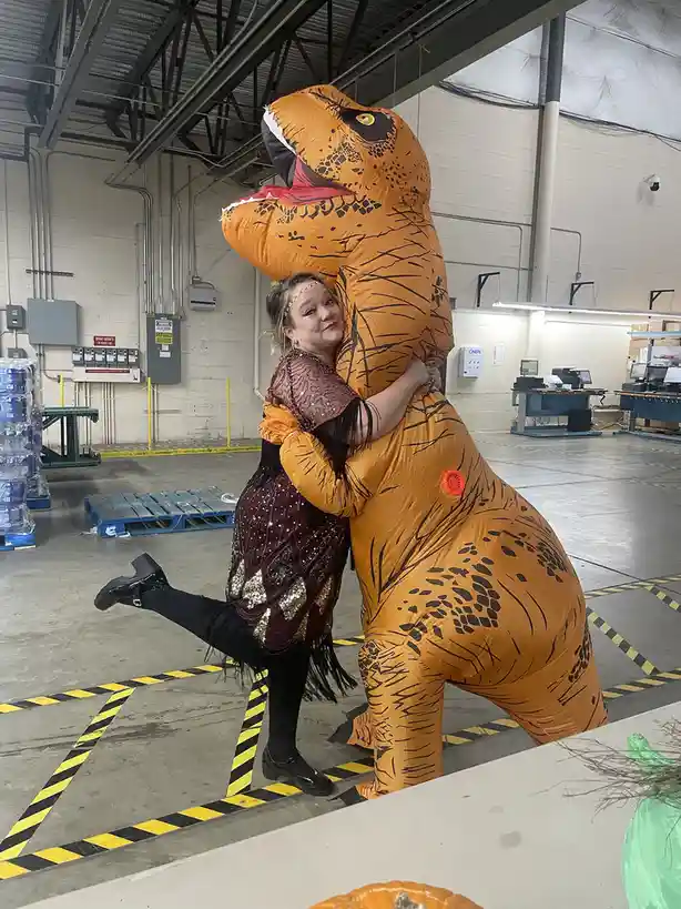A woman in a sparkly, sequined costume hugs a large, orange inflatable T-Rex costume in a warehouse setting. She has one leg…