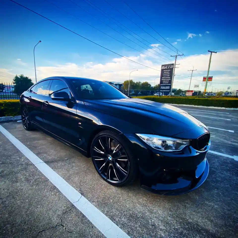 A glossy black BMW 330i sedan is parked in a commercial parking lot under a bright blue sky with scattered clouds. The car…