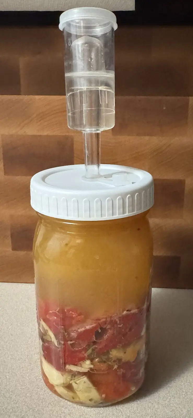 A glass jar filled with chopped red and yellow vegetables submerged in brine, topped with a white lid and a clear airlock…