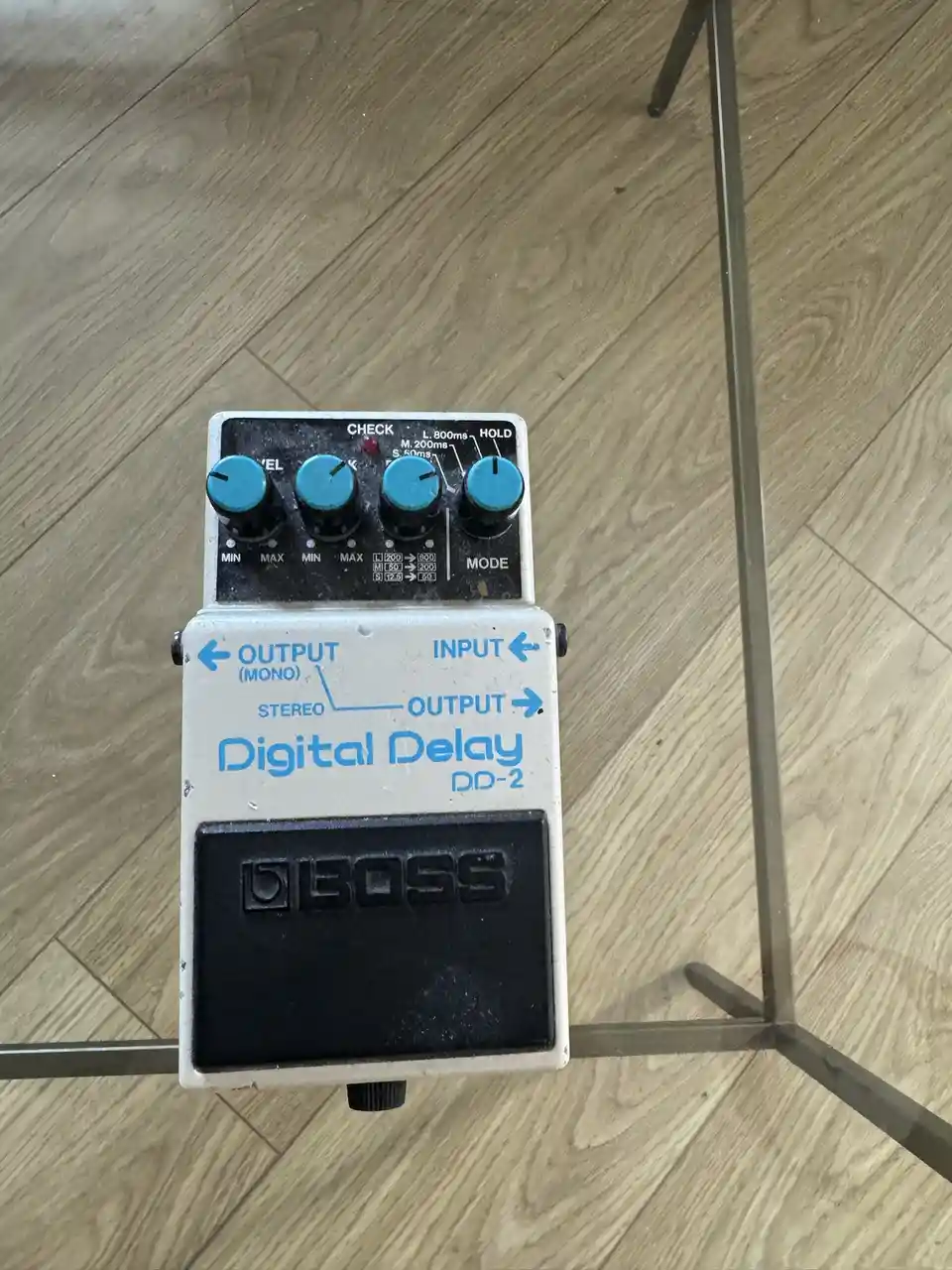 A Boss DD-2 Digital Delay guitar effects pedal with four blue knobs and a black footswitch rests on a light wood floor…