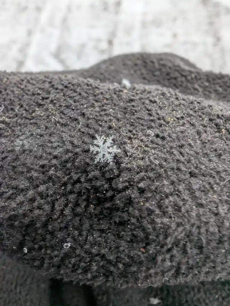 A close-up photo shows a single, intricate snowflake resting on the textured, dark gray fabric of a knit garment, with a…