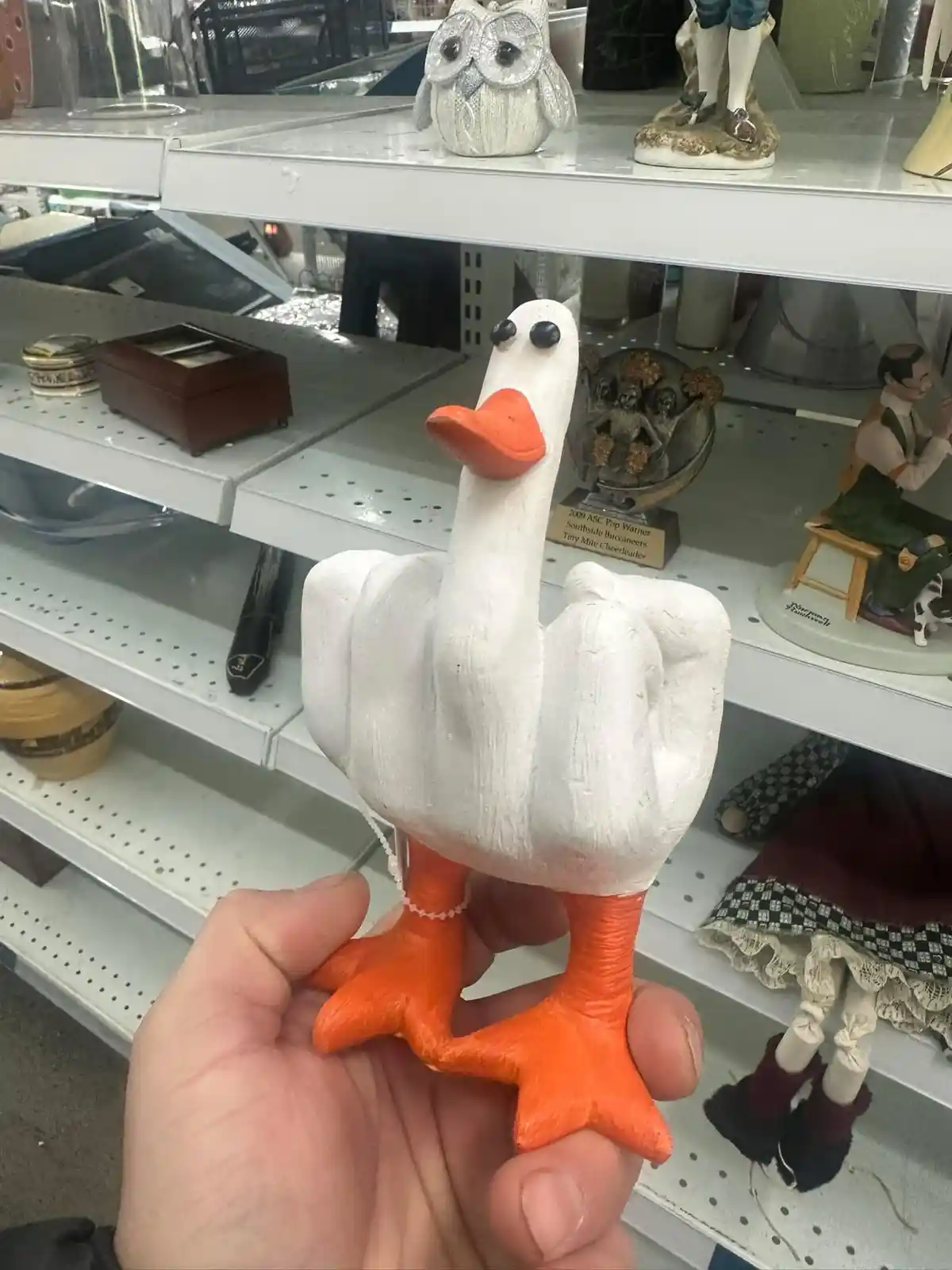 A hand holds a white ceramic goose figurine with an orange beak and feet, standing on a store shelf among other decorative…