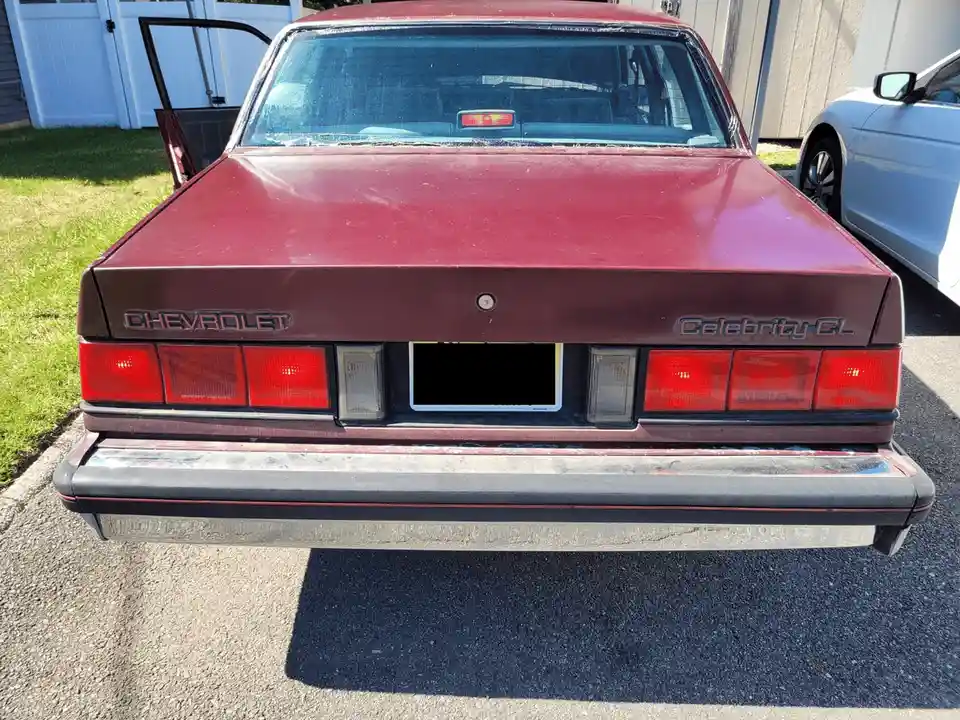 The rear view of a maroon 1980s Chevrolet Celebrity CL sedan parked outdoors, showing its taillights, 'CHEVEY' and…