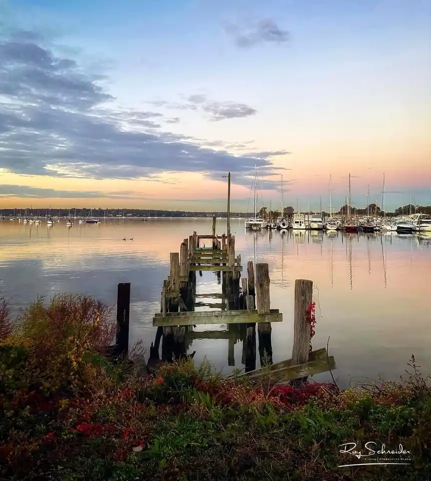 A serene sunset photo of a weathered wooden pier extending into calm water, with sailboats docked in the distance and autumn…
