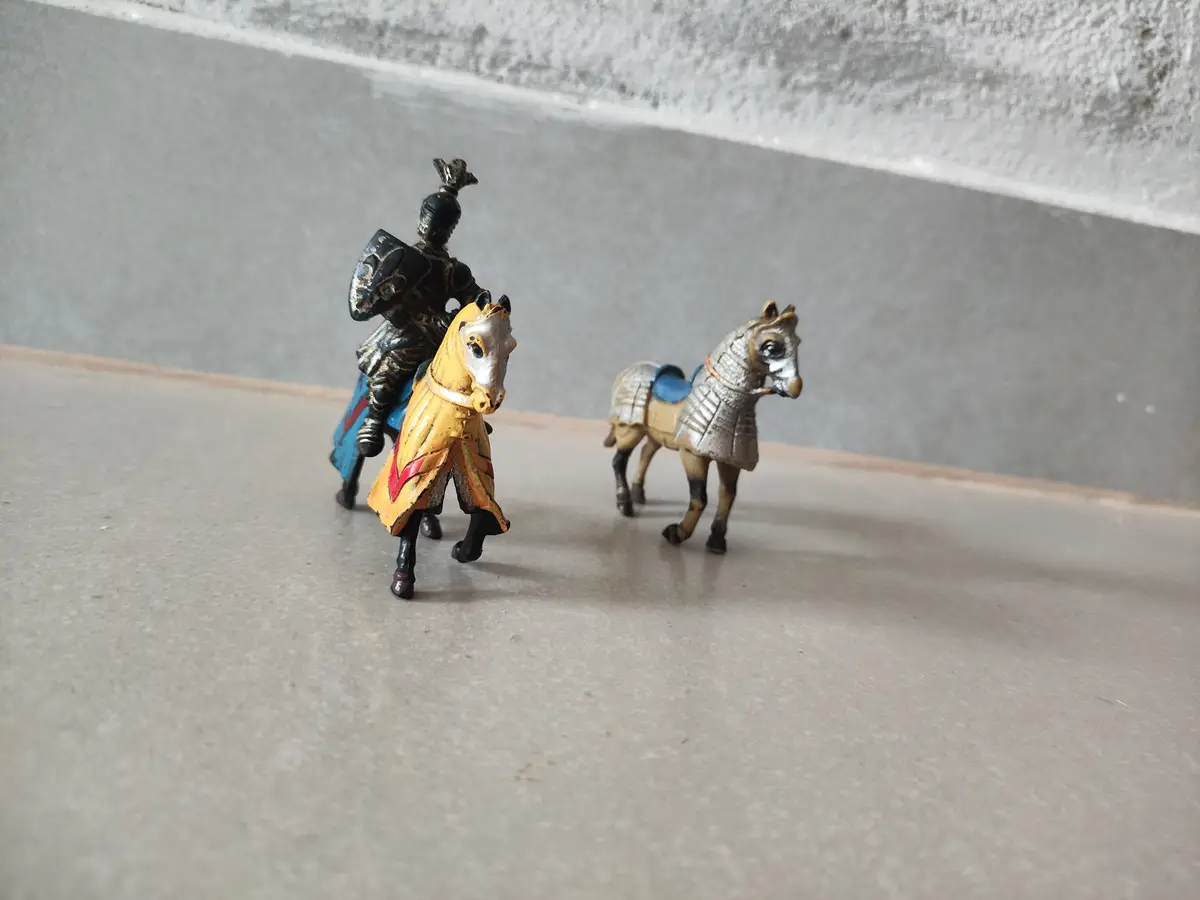 A toy knight figurine on a horse stands next to a separate toy horse, both on a plain surface. The knight is holding a…