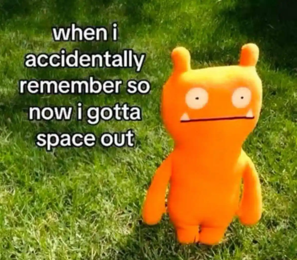 An orange Uglydoll plush toy with a blank, unblinking stare stands on green grass. White text with a black outline is…