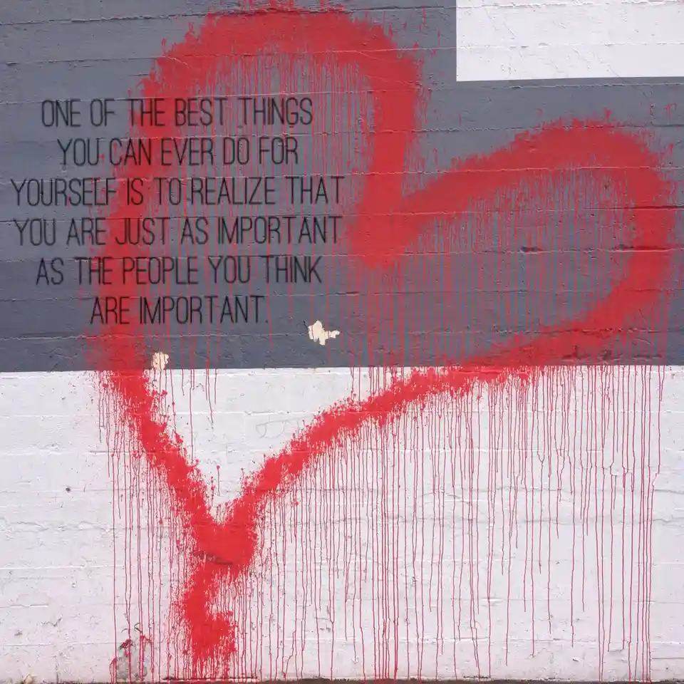 A mural on a brick wall painted with a red heart and dripping red paint over a gray and white background. Black text…